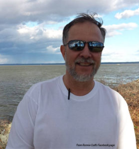 A white male with a graying beard is standing in front of what appears to be a lake, smiling . He has on a white t-shirt and mirrored sunglasses that show the reflection of someone taking his photograph. Under his t-shirt is a pen clipped to his collar.