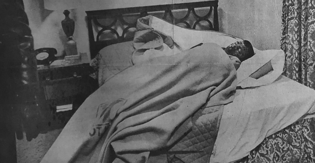 A black and white photo of Charles Bright wrapped up in blankets on a hotel bed after his fall.