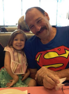 Charles Bright with his granddaughter A balding white man smiles as he wears a Superman t-shirt. To his right is a little girl, his granddaughter.