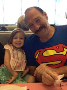 A balding white man smiles as he wears a Superman t-shirt. To his right is a little girl, his granddaughter. 