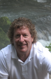 A brown curly-haired man sits in front of a moving river.