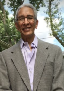 A brown-skinned man in a suit jacket and purple button-down shirt is smiling, his hair gray, in front of a blue cloud-filled sky and trees.