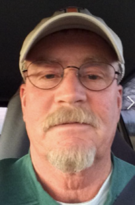 A white man is in his car, looking at the camera. He has a blonde/grayish goatee, glasses, and a baseball cap on.