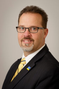 A light-skinned man with black-rimmed glasses and a salt-and-pepper goatee smiles into the camera. He is wearing a suit and a PSD pin.