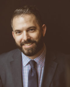A light-skinned, bearded man with brown hair is in a suit, smiling at the camera.